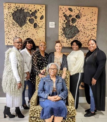Seven women gathered around a seated elder in a leopard-print armchair